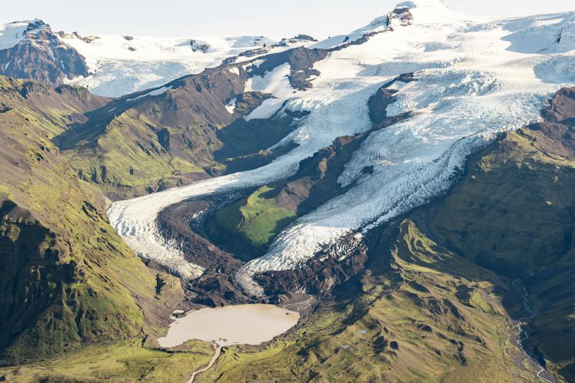 An aerial view of a broad glacier flowing between steep green and dark brown mountain slopes, ending in a large pale meltwater lagoon on a valley floor.
