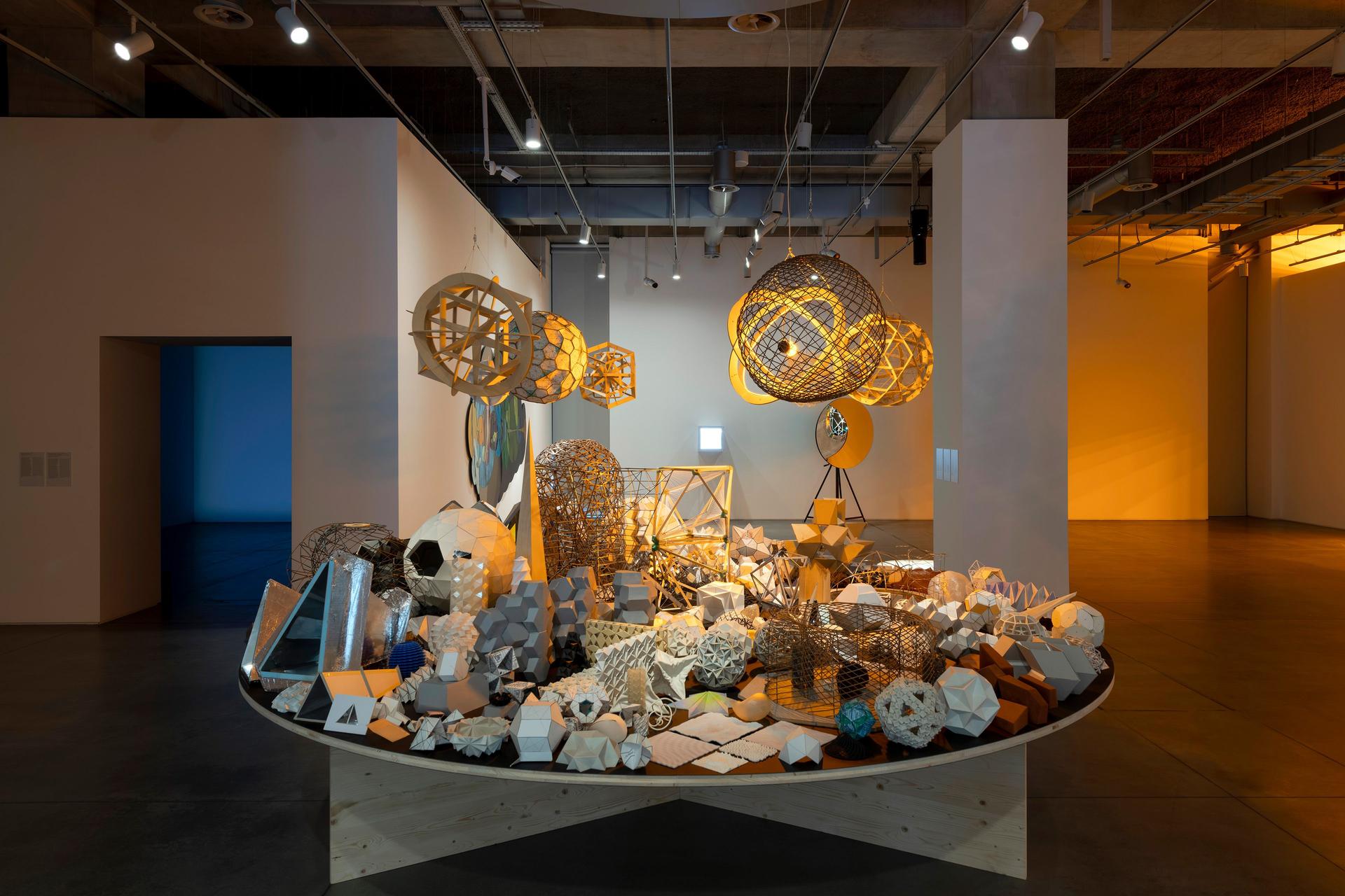 A large round table densely in a dimly lit gallery space, filled with geometric models and sculptural forms, with several illuminated spherical structures hanging above.