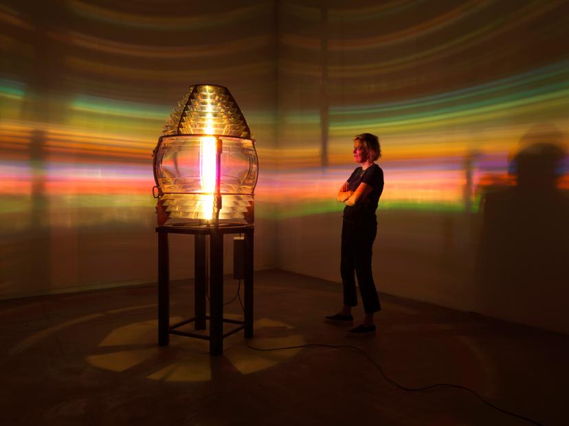An illuminated lens mounted on a wooden plinth in an otherwise darkened gallery, casting circular rays of coloured light across the gallery walls. A visitor stands nearby, arms crossed, surrounded by bands of refracted rainbow light spreading across the walls.