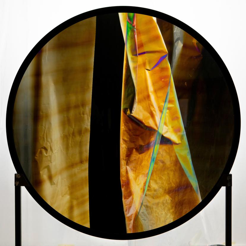 large circular frame encloses overlapping yellow, black and multicoloured translucent sheets, with visible creases and soft reflections.