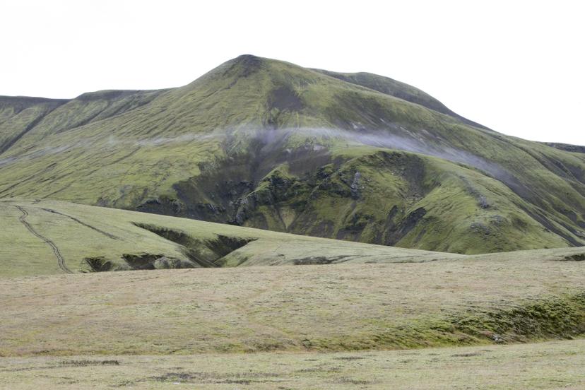 A thin misty cloud drifts across a green mountain landscape, with dark fissures and gentle slopes across the surrounding volcanic terrain.