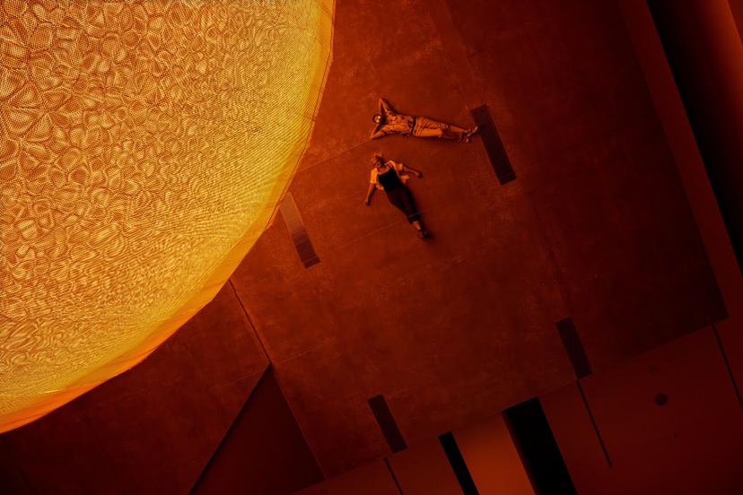 Viewed from above, two people lie on the gallery floor beneath a glowing yellow spherical artwork, which enters the upper frame and casts warm light across the surrounding space and mirrored surfaces.