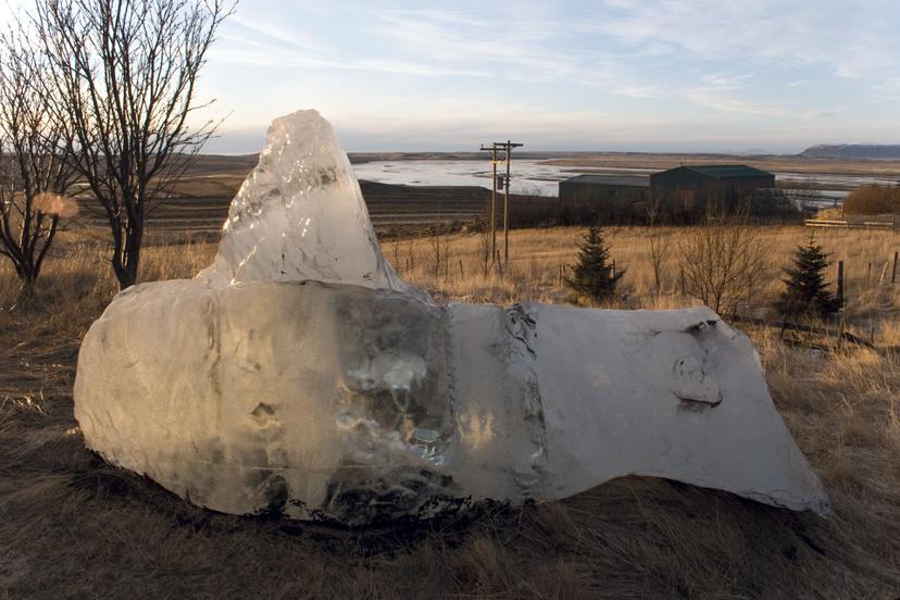 A large block of ice sits on dry grassland under pale afternoon light. The transparent form glows faintly against a backdrop of bare trees, farm buildings, and a distant river plain.