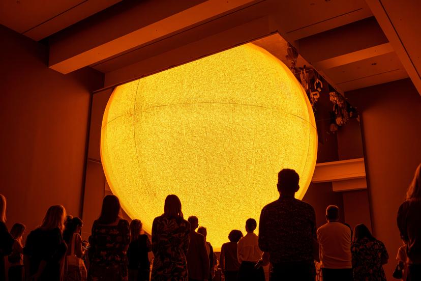 A crowd of silhouetted visitors stand in a darkened gallery looking up at a large glowing sphere suspended overhead, its bright yellow surface covered in a dense pattern of small curved forms that create a moiré effect.