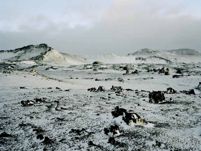 A snow-covered volcanic plain scattered with dark, irregular rock formations, with low ridgelines and distant slopes rising beneath an overcast sky.