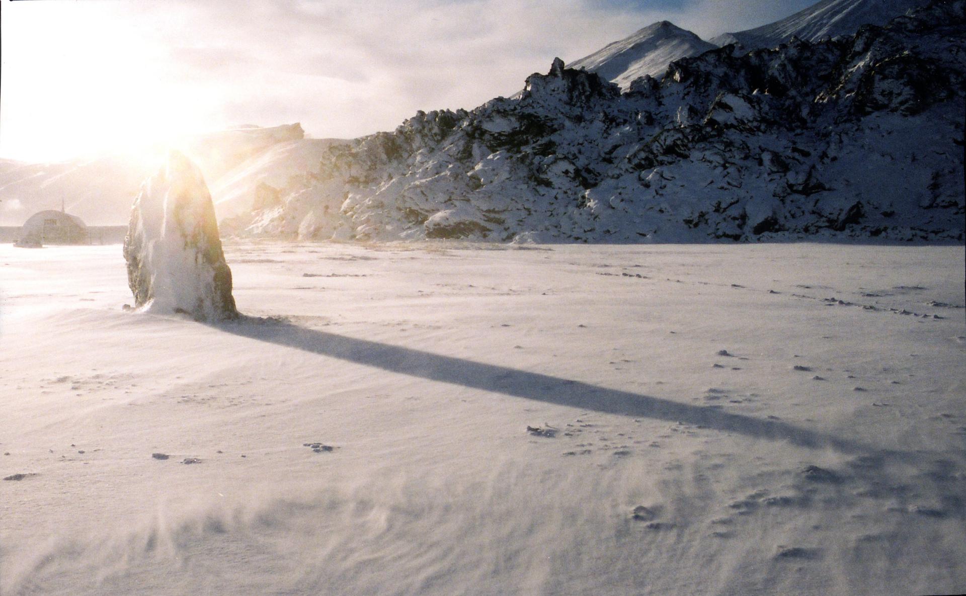 A tall, snow-covered rock stands in a wide, frozen landscape with a long shadow stretching across the snow, illuminated by low sunlight against a backdrop of rugged, snow-covered mountains and a dome-shaped structure.