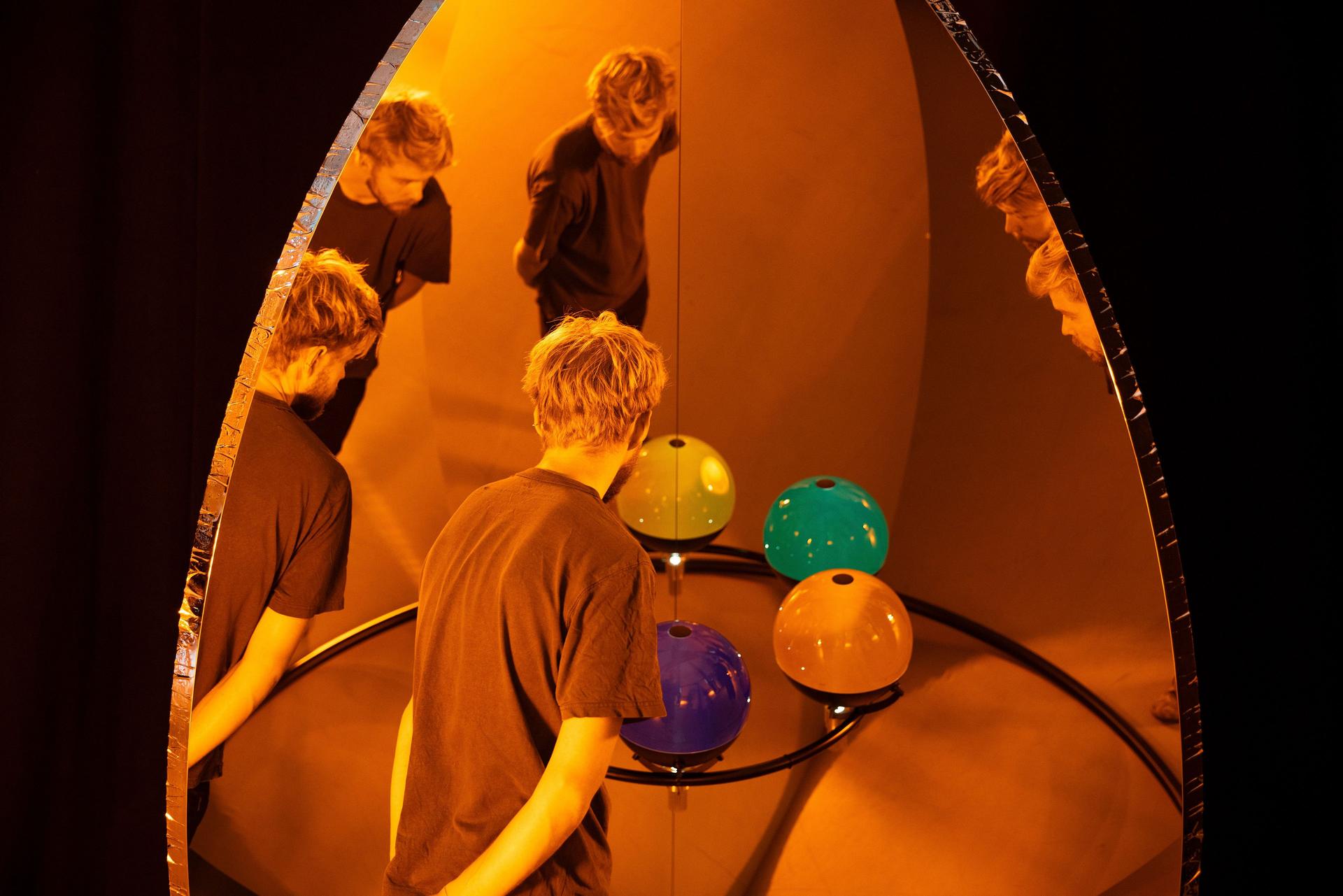 A person leaning over a circular frame holding glossy coloured spheres, which appear blue, teal, green and amber, while multiple reflections of their figure appear in the tall mirror behind the arrangement.