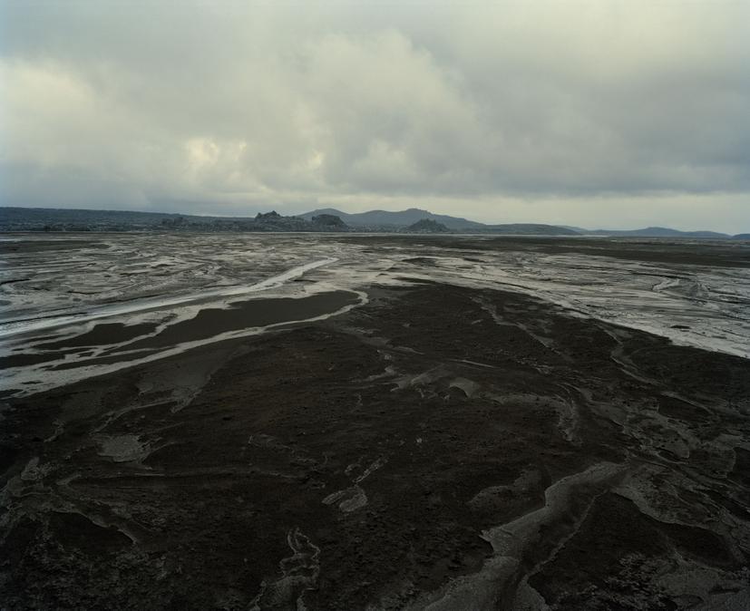 A broad expanse of dark, rippled earth and silt extends toward a low, rounded hill under a cloudy sky, with multitudinous thin water channels and scattered grey patches visible across the barren, wetland terrain.