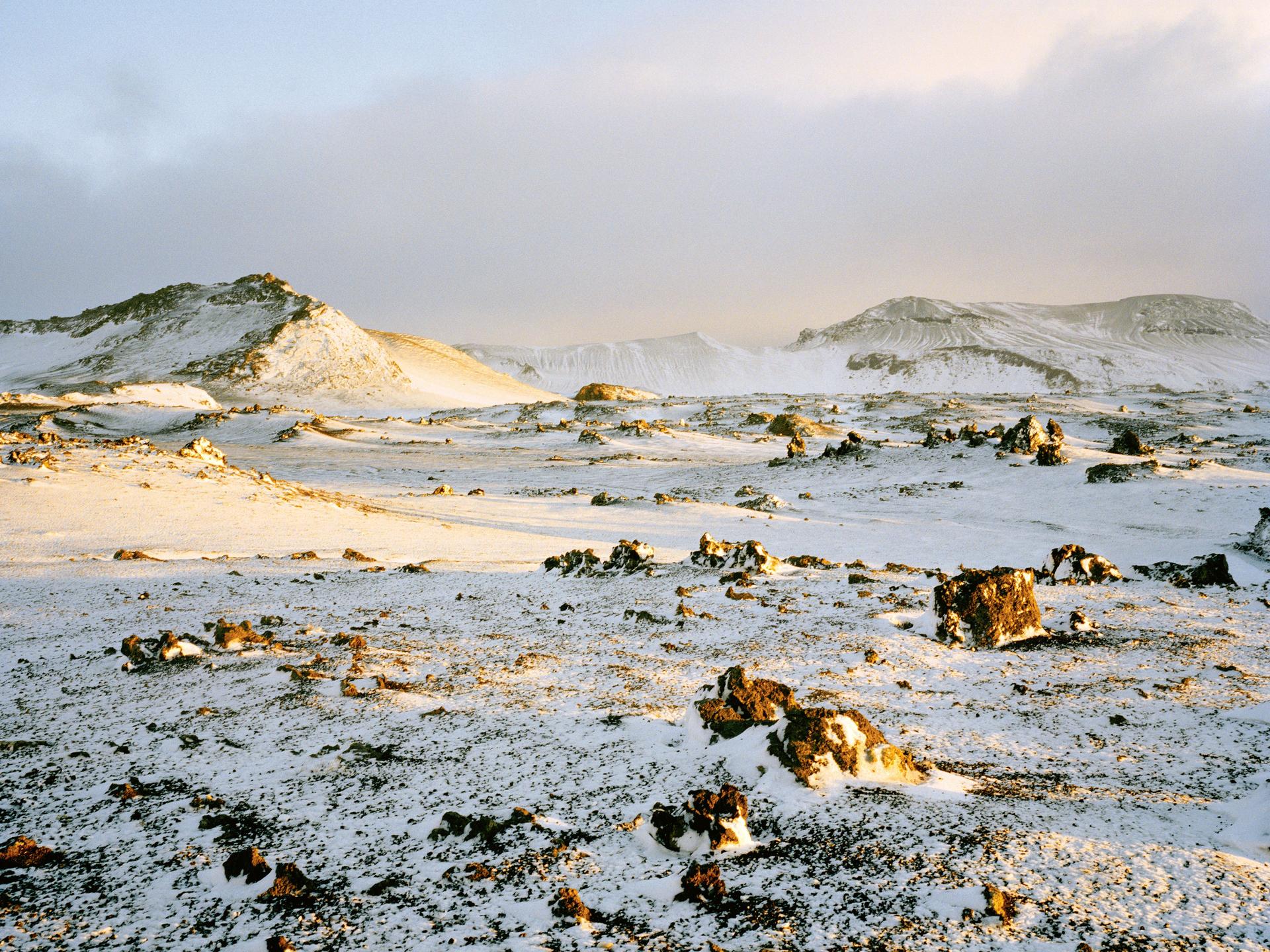 A snow-covered volcanic plain dotted with dark, irregular rocks, lit by warm yellow light, with low ridges and distant slopes under a cloudy sky.