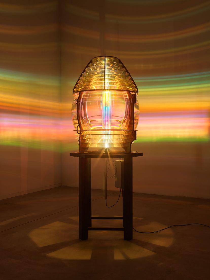 An illuminated lens in an otherwise darkened space, casting circular rays of coloured light across the gallery walls.