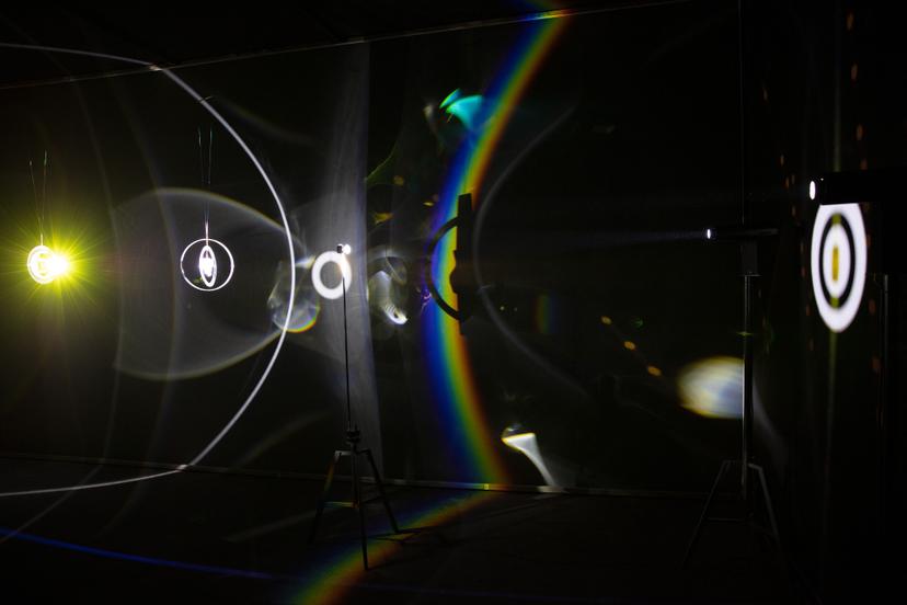 A dark room is lit with overlapping lights in bright rings, thin white arcs and blurred sheets across the walls and floor. Lights on tripod stands cast beams that refract into sweeping curves and dispersed colours, including a vivid rainbow band, while suspended circular objects create sharp halo-like outlines and scattered reflections form small glowing points.