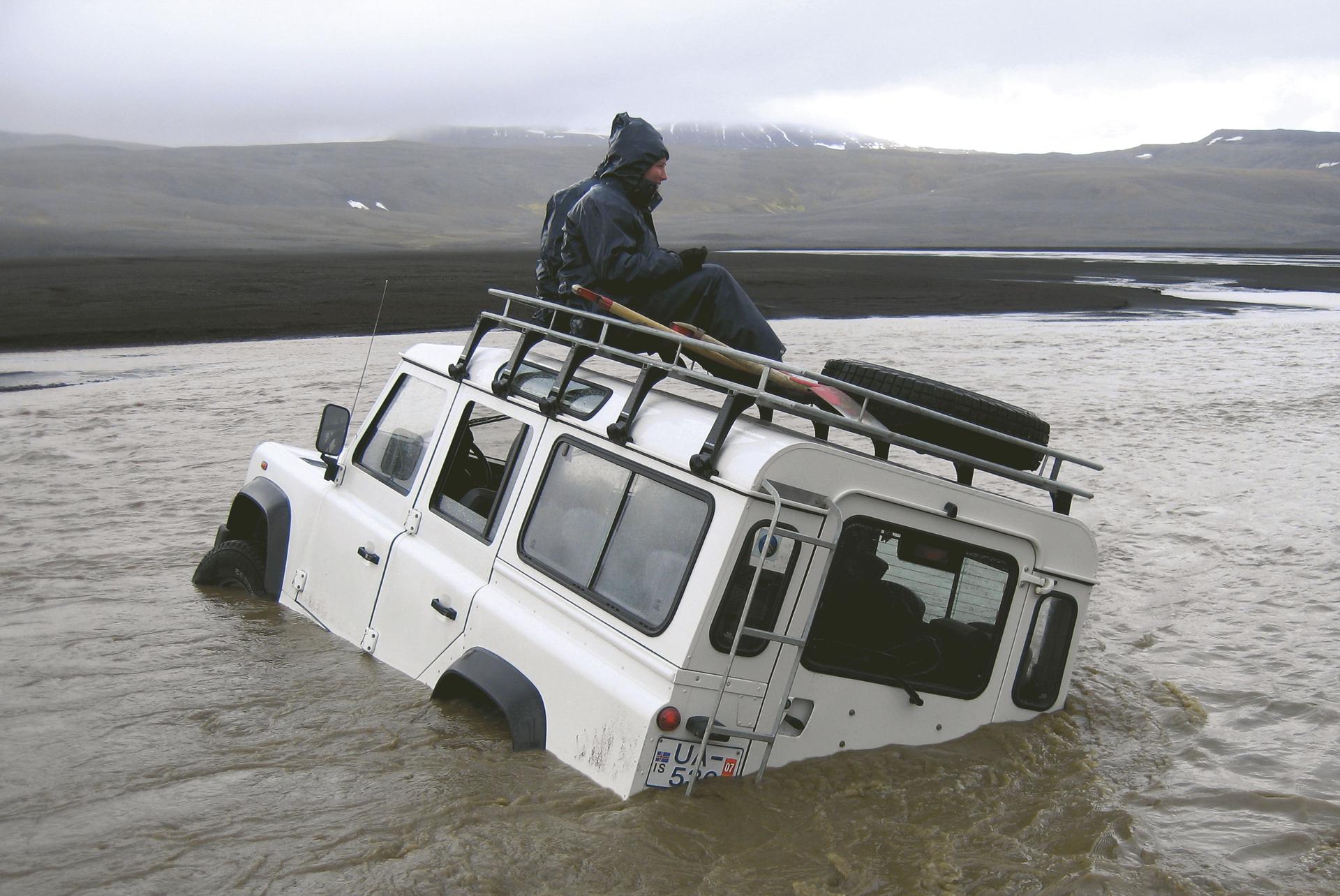 A person in a hooded rain jacket sits on the roof rack of a white off-road vehicle tilted and sunk in moving brown water, with dark hills in the distance.