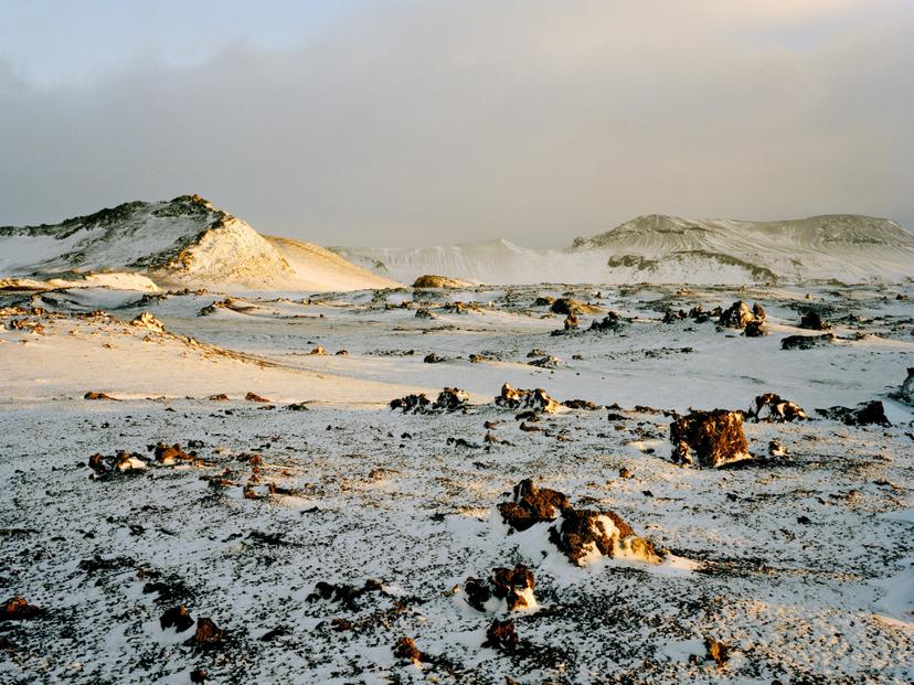 A snow-covered volcanic plain scattered with dark, irregular rocks, lit by warm yellow light, with low ridgelines and distant slopes beneath a clouded sky.