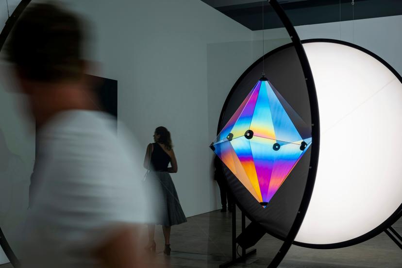 A blurred passer-by moves in front of a large circular frame containing a faceted geometric form glowing from within, its translucent panels blending blue, yellow and pink as two visitors observe from behind.