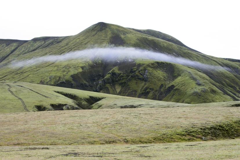 A wide landscape with a misty band of cloud suspended mid-slope above a green mountain, under the diffuse light of a white sky.