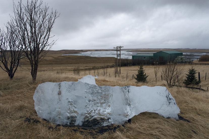 The ice has partially melted, revealing a thin reflective layer on the ground. The surrounding field stretches toward a river and low hills beneath a soft, blue-grey sky.