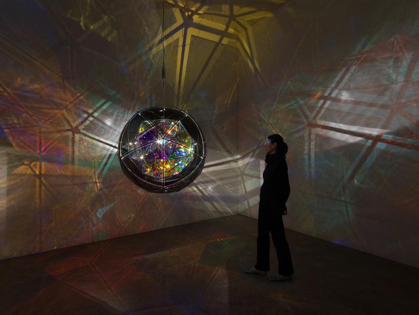 A person stands in a darkened gallery facing a large, faceted spherical structure that glows with iridescent, multicoloured light, reflecting geometric patterns across the gallery walls.