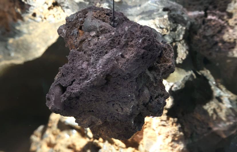 A dark, irregular rock with porous cavities and rough ridges hangs on a thin black cord against a blurred backdrop of pale, uneven surfaces and shadowed hollows.