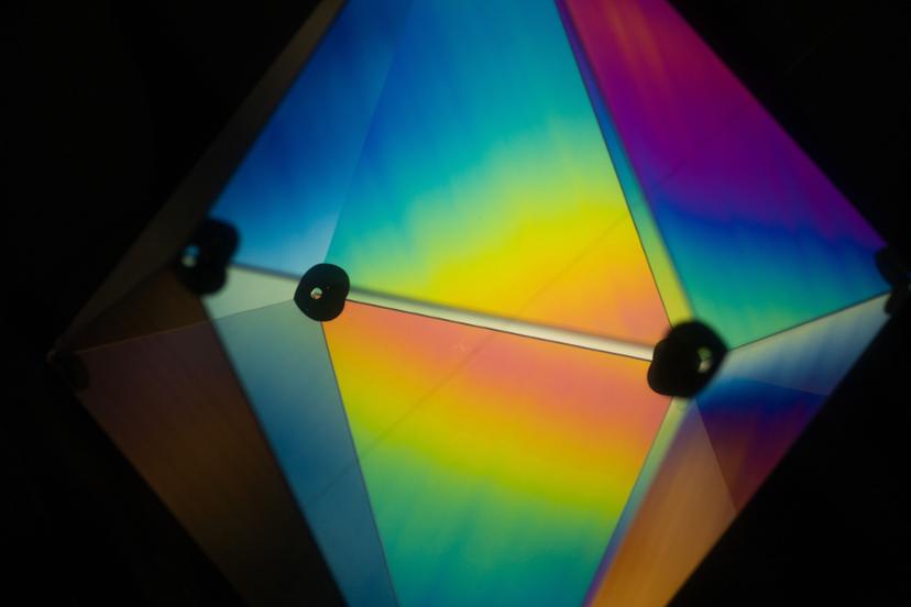 A faceted geometric structure glows from within, its translucent panels showing smooth gradients of yellow, blue, pink and violet around dark circular connectors.