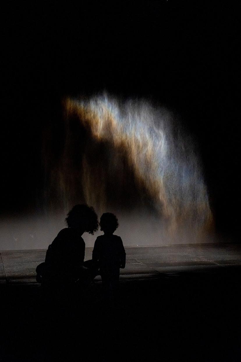The silhouettes of an adult and child stand before a curtain of mist, partially illuminated by light, forming a glowing multicoloured arc across the water spray in an otherwise darkened space.