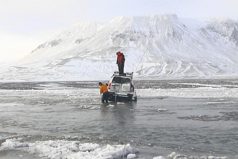 Two people stand near a white off-road vehicle in shallow icy water. One person stands on the vehicle’s roof and the other is wading near the wheel, with snow-covered mountains across the horizon behind them.
