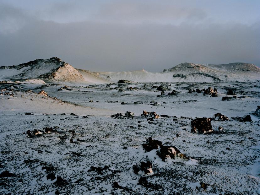 A snow-covered volcanic plain scattered with dark, irregular rocks, with low ridges and distant slopes beneath a dim, cloudy, blue-grey sky.