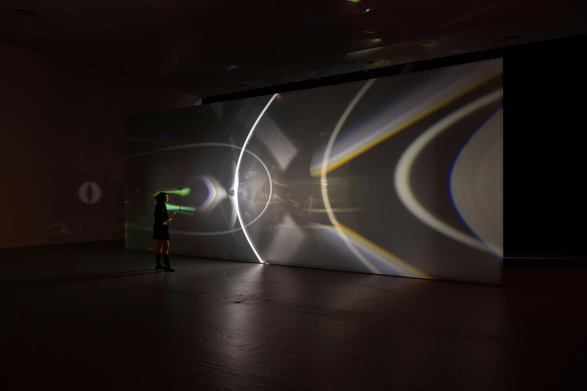 A person stands in a spacious dark gallery facing a tall white wall covered in sweeping bands and arcs of bright light and rainbow colours, including broad white curves and dispersing yellow and blue streaks that extend across the wall’s length.