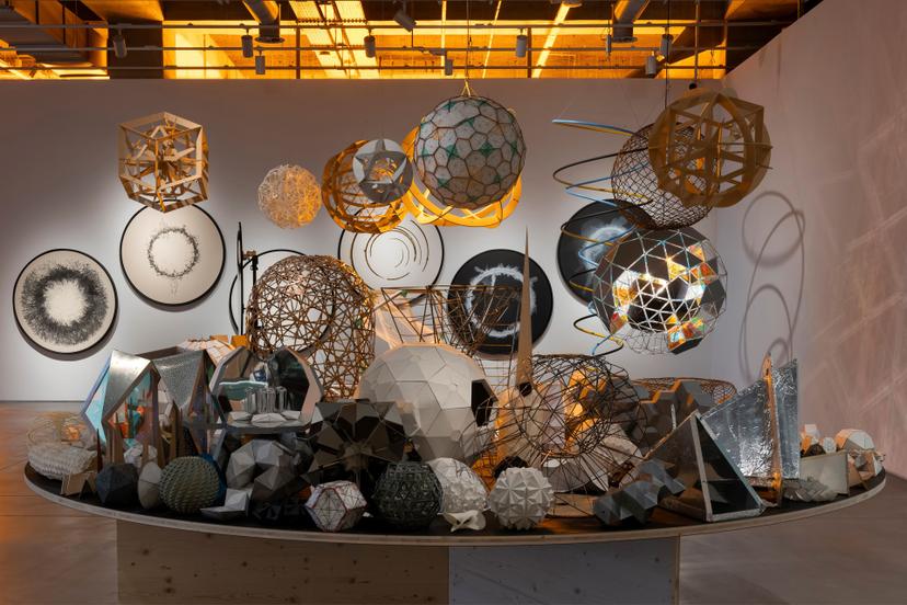 A round table densely packed with geometric sculptures in various materials, with illuminated spherical structures hanging above and circular artworks on the wall behind, featuring dense, ring-shaped clusters of black, ink-like marks.