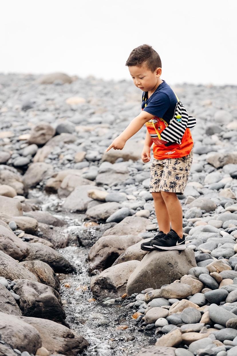 A young child stands on a large rock beside a narrow stream of water flowing through grey and tan stones, leaning forward and pointing towards the moving water in a white-walled gallery.
