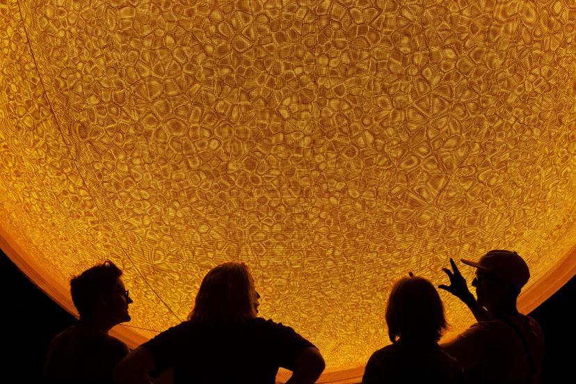 Silhouetted visitors stand below a massive yellow glowing sphere whose surface shows densely arranged curved forms, creating a dynamic moiré pattern as people look upward.