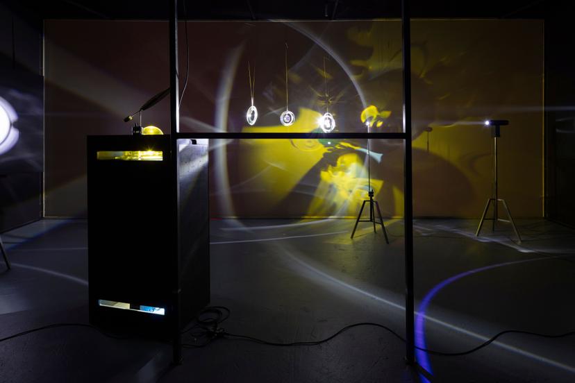 A dark room contains several spotlights and hanging circular objects that cast overlapping projections of bright rings, sweeping white arcs and broad yellow washes across the walls and floor. A tall black box with illuminated openings stands in the foreground beside metal poles and trailing cables, while tripod-mounted lights create additional curved bands, soft flares and shifting reflections throughout the space.
