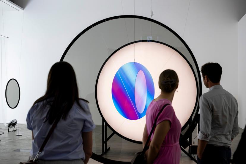 Three visitors stand before a circular frame holding a glowing geometric form that displays smooth colour gradients of blue, purple and pink across its translucent panels, joined by dark circular connectors.