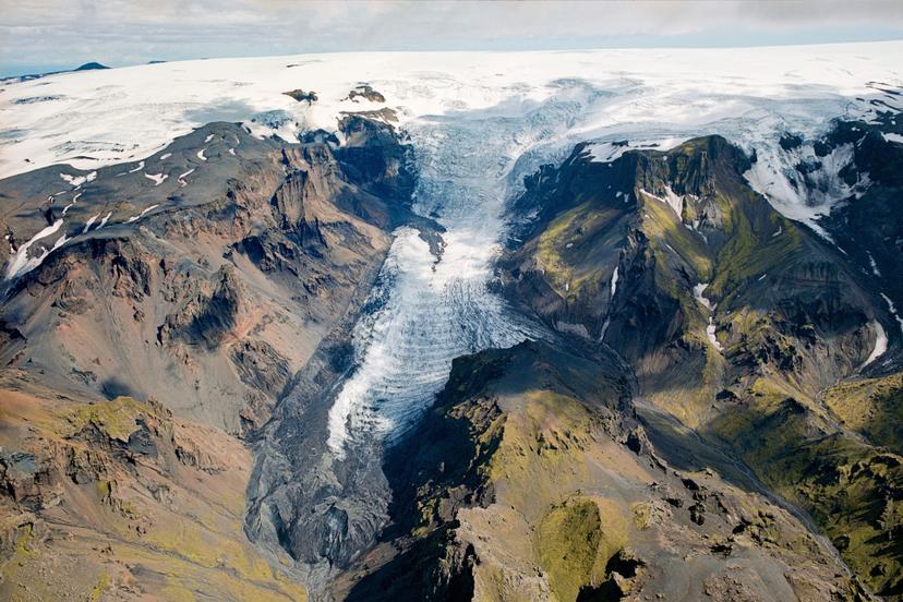 An aerial view of a glacier descending steeply between steep mountain ridges, its white and blue ice bordered by snow, dark rocky slopes and patches of green moss.