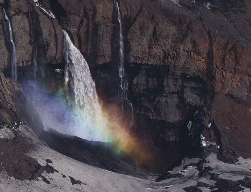 A waterfall cascades down a rocky cliff face, with a vivid rainbow mist hovering above the surface of the water, set against dark volcanic stone.