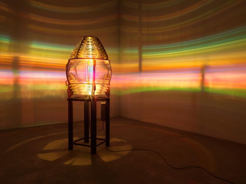 A glass lens glowing with a vertical column of rainbow light. The surrounding space is bathed in multicoloured illumination.