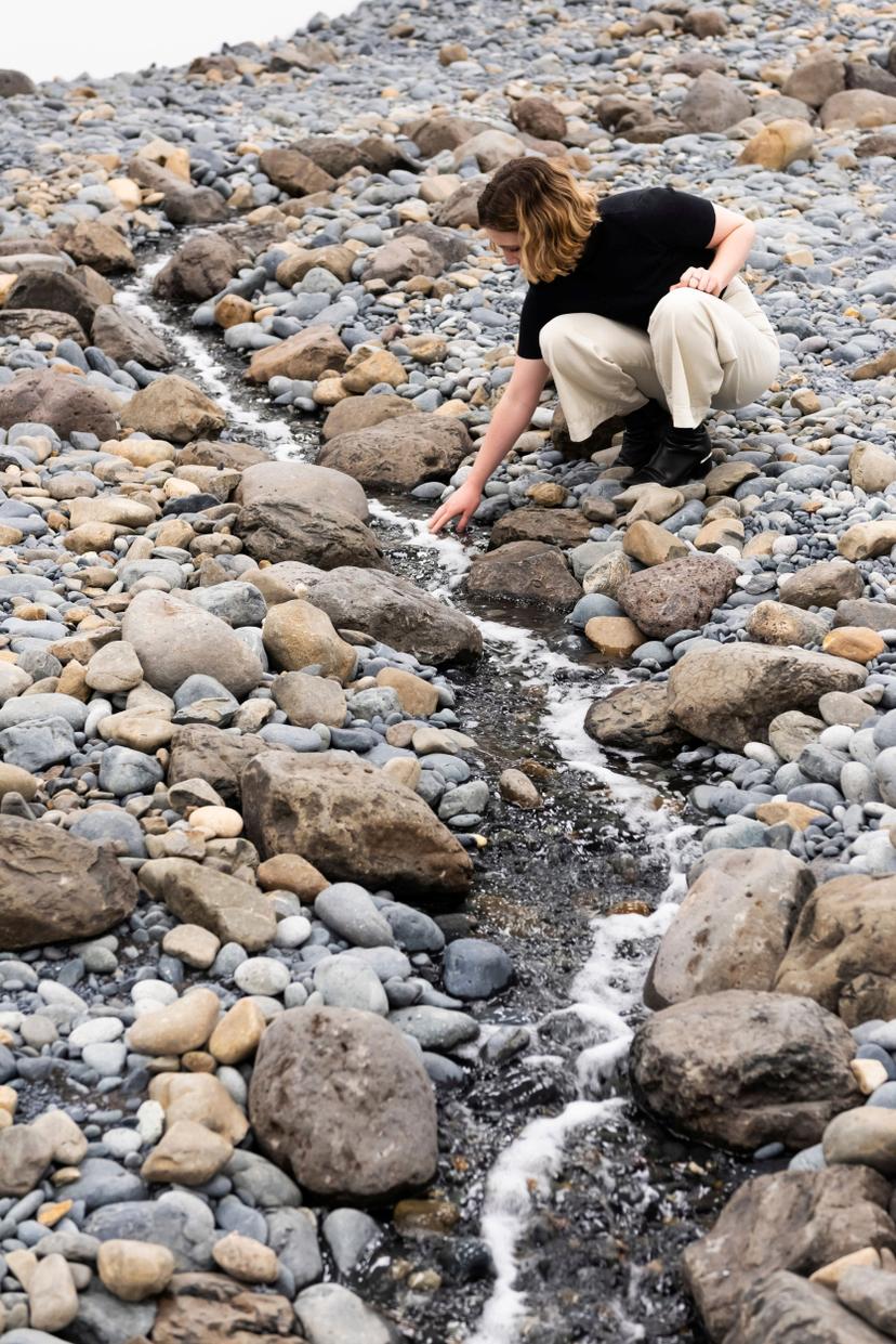 A person crouches on a rocky surface and extends a hand into a thin stream of running water that winds between grey and tan stones in a white-walled gallery.