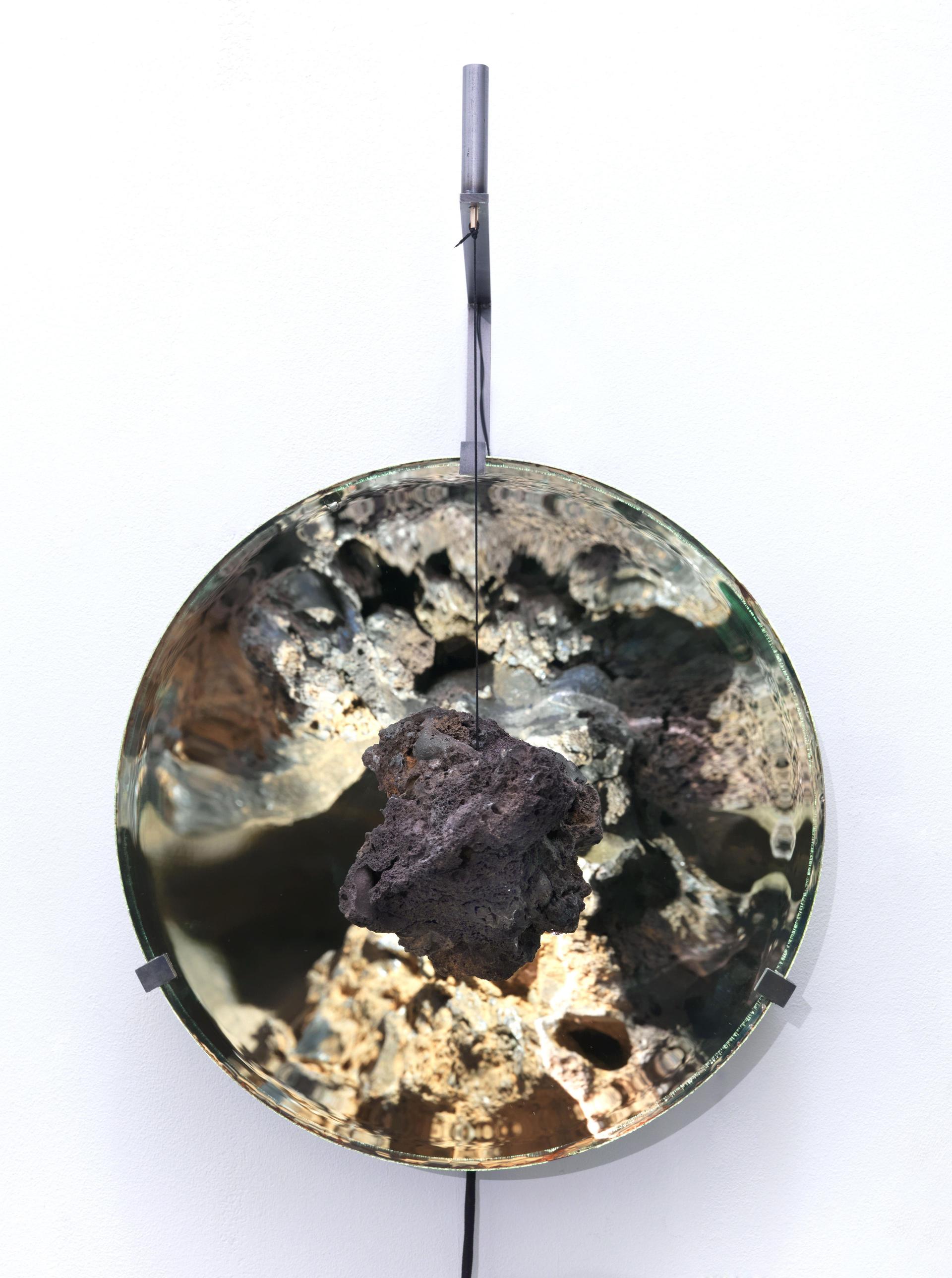 A dark, porous stone hangs from a thin black cord in front of a large concave mirrored disc, whose rippled metallic surface reflects distorted light and crater-like textures, with the disc held by small metal clips and suspended from a vertical metal bracket on a plain white wall.